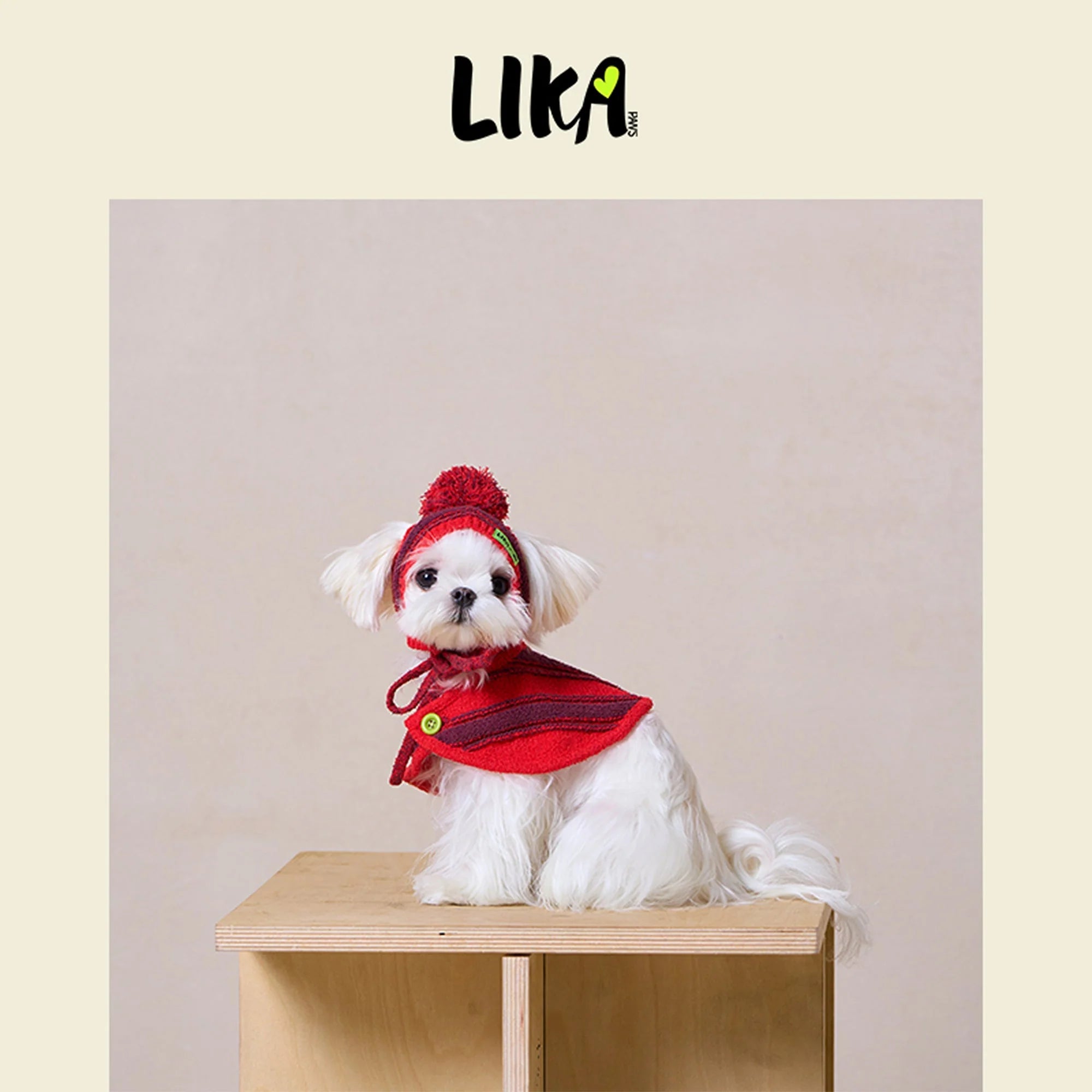 Small white dog wearing a red outfit with a hat on a wooden table against a beige background, featuring the LIKA brand.