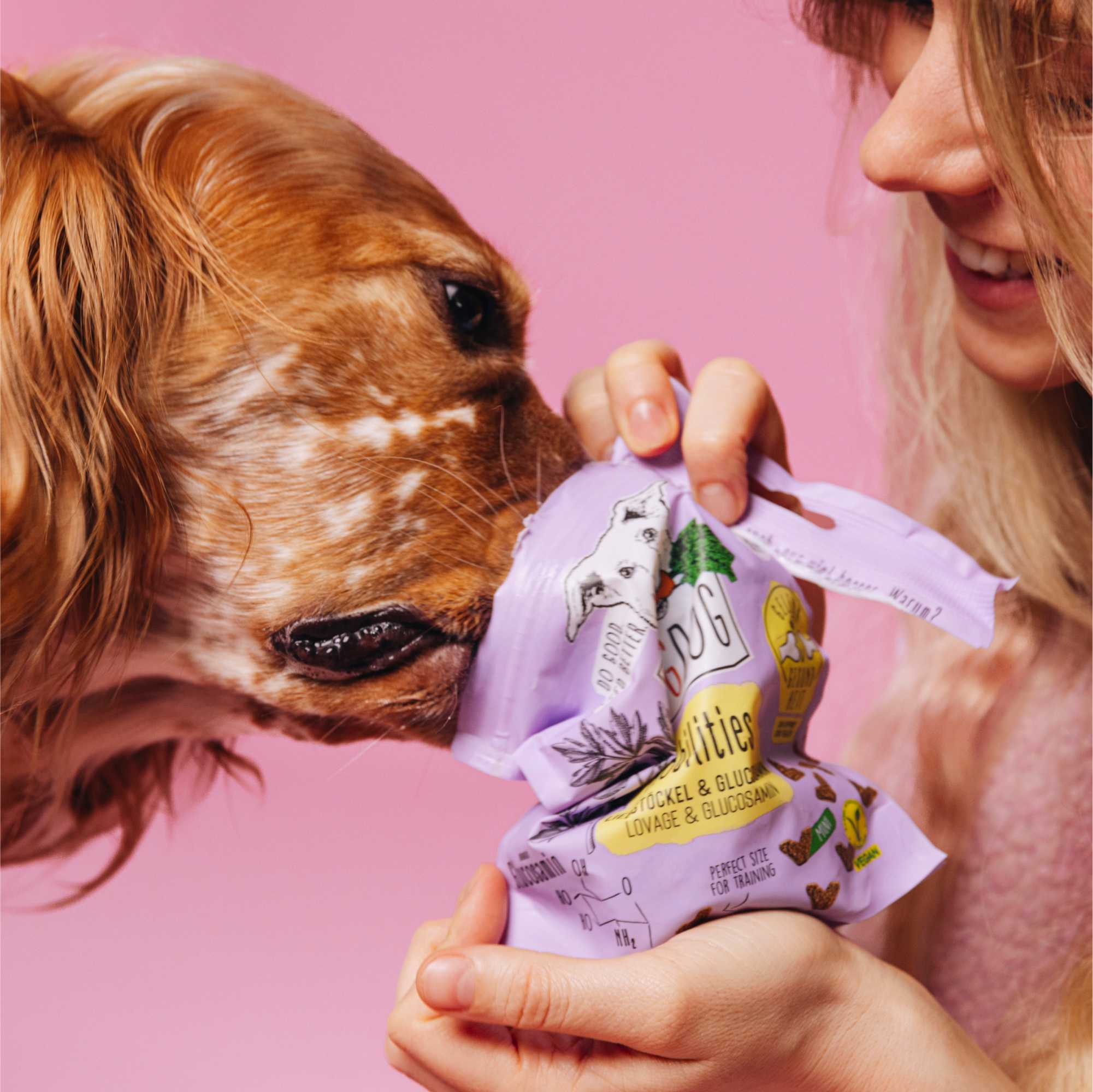 Woman holding a pet product package with a dog sniffing it against a pink background
