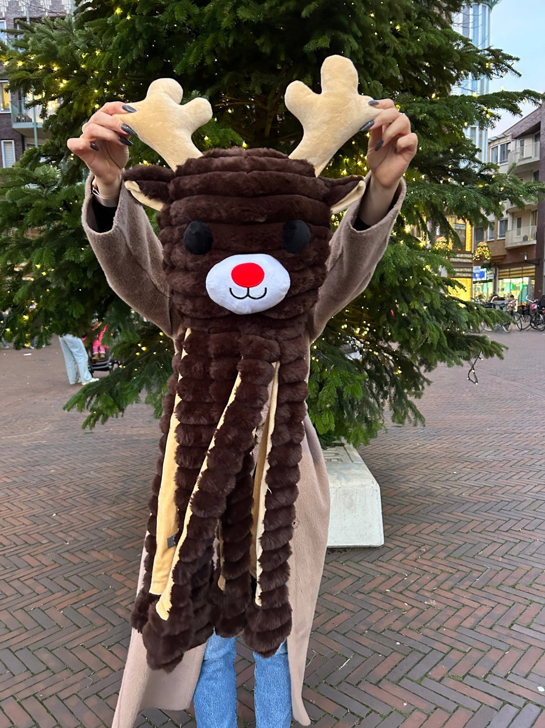 Person wearing a reindeer costume in an outdoor setting with trees and buildings in the background.